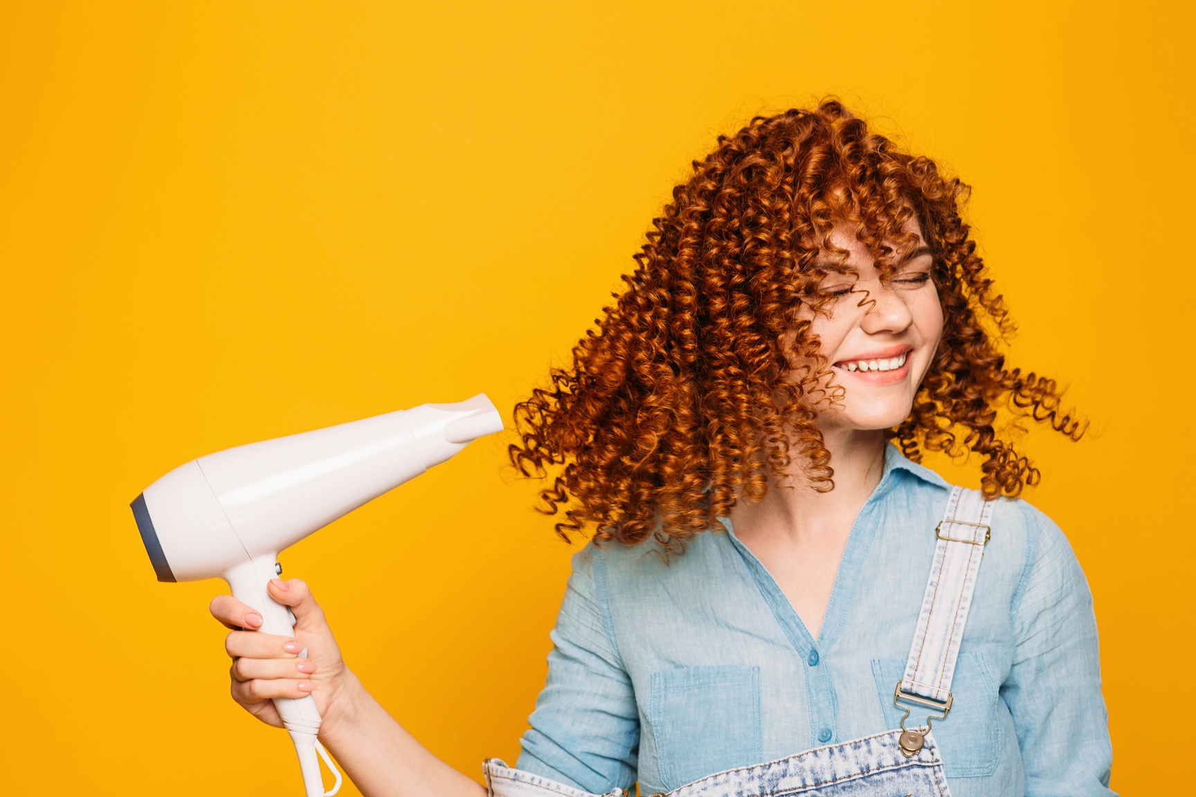 curly red-haired woman using hair dryer on yellow background. Making perfect curls